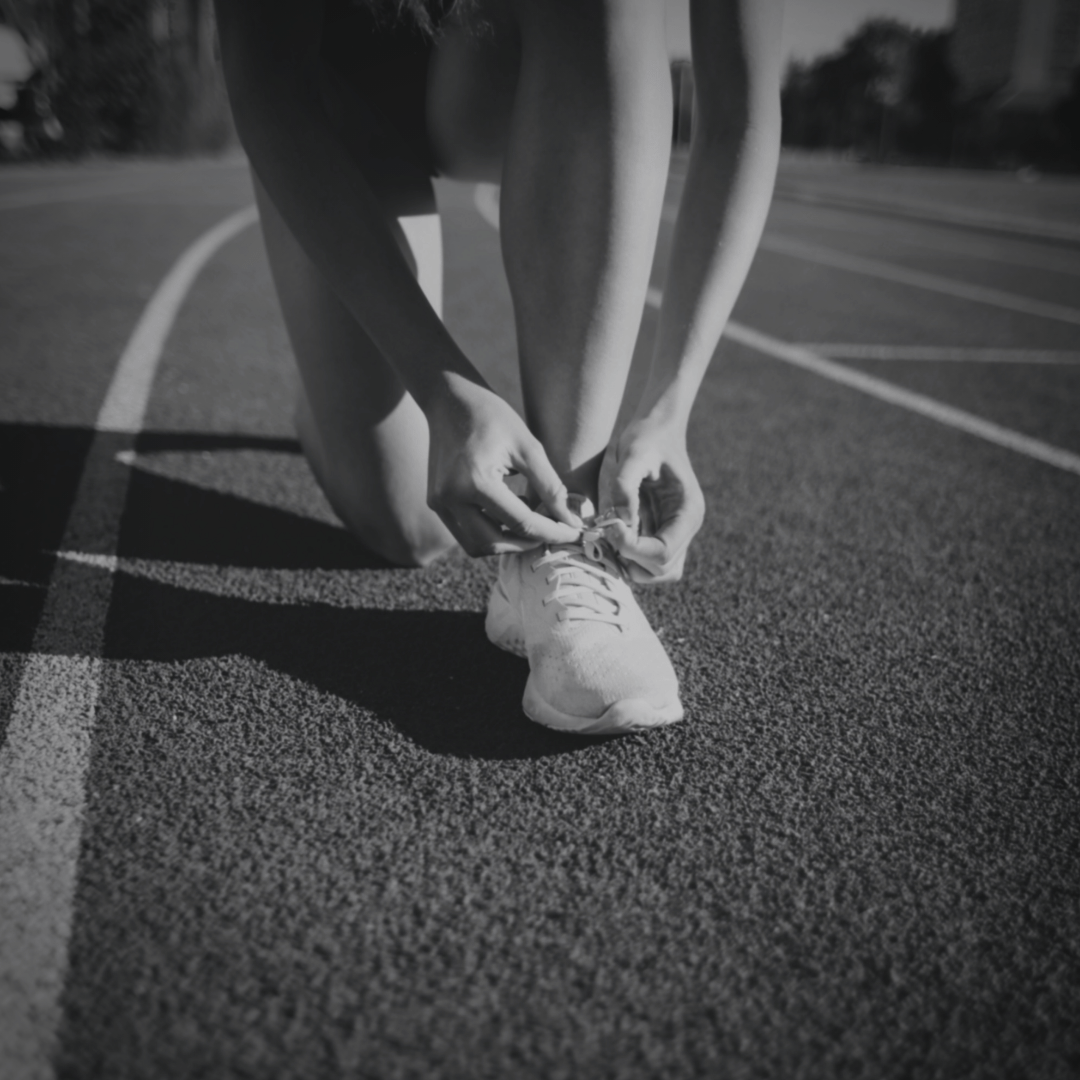 A person kneels on a running track, tying the laces of a white athletic shoe, preparing for a workout or race.