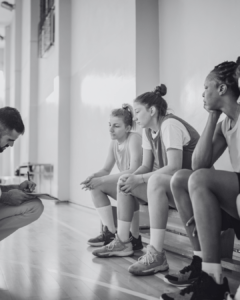 A coach discusses strategy with three seated female basketball players, emphasizing teamwork and preparation in a gymnasium setting.