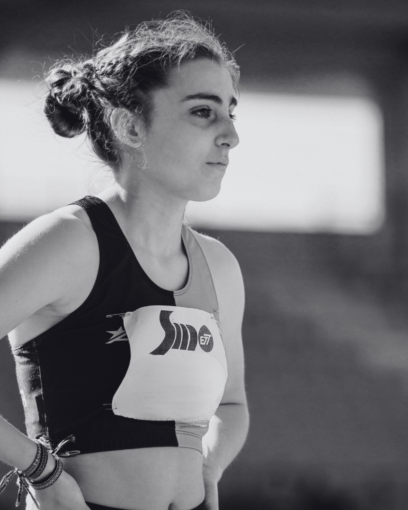 Teenage female athlete focused and poised on the track, wearing a black sports top and competition bib, with sunlight highlighting her determination.