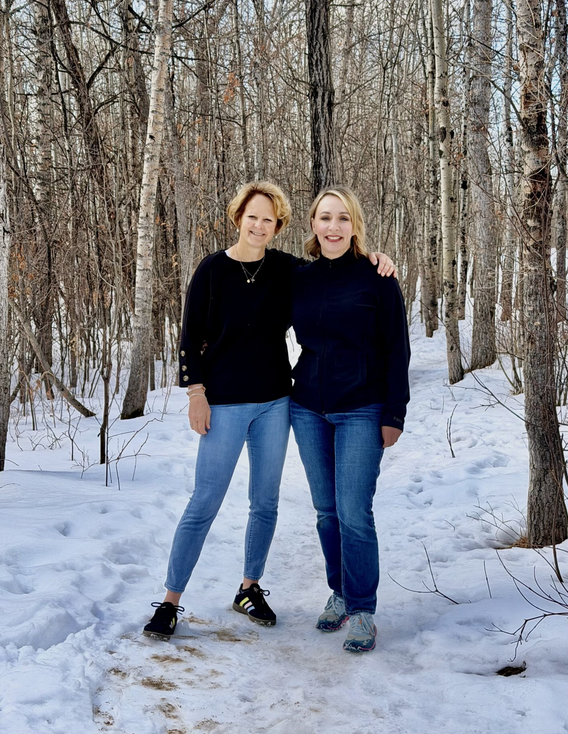 Two women stand together smiling on a snow-covered path in a forest. The scene captures a moment of friendship in a winter setting.
